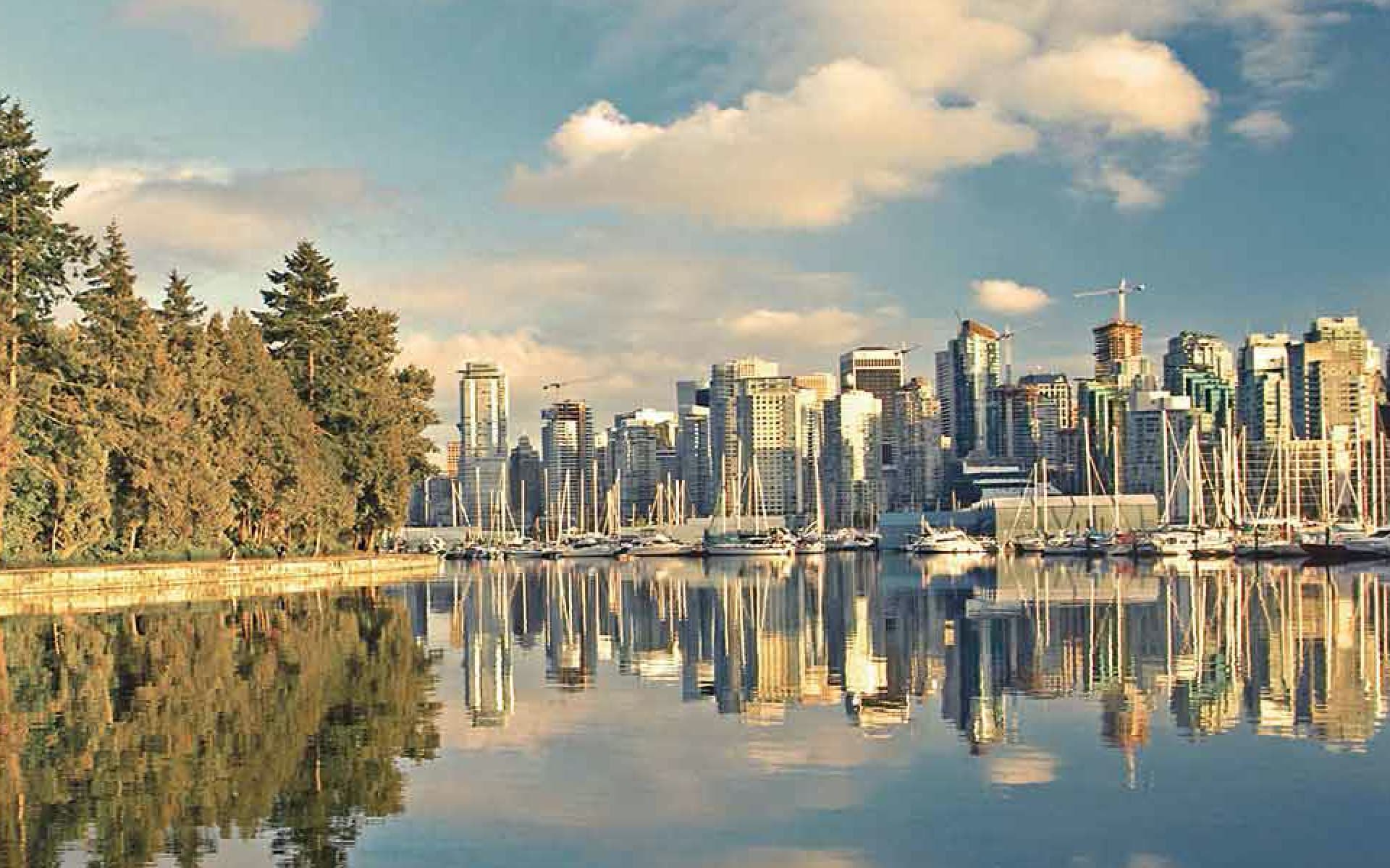 A picturesque view of the Vancouver, Canada skyline, featuring its towering skyscrapers reflecting in the calm harbor waters, bordered by lush trees and a fleet of sailboats.
