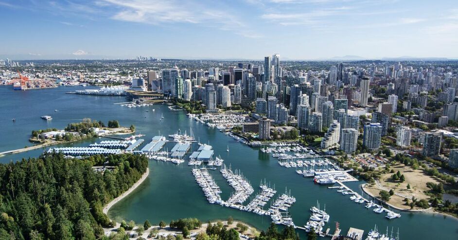 This is an aerial view of a bustling city harbor, with numerous boats docked and a dense skyline of skyscrapers. Green forested areas are visible on the left, contrasting with the urban landscape.