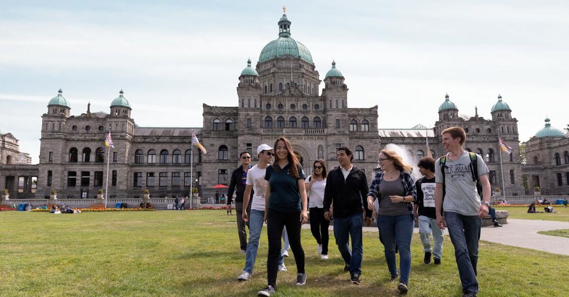 A group of tourists strolls across a manicured lawn in front of a grand, historic building featuring a prominent dome.