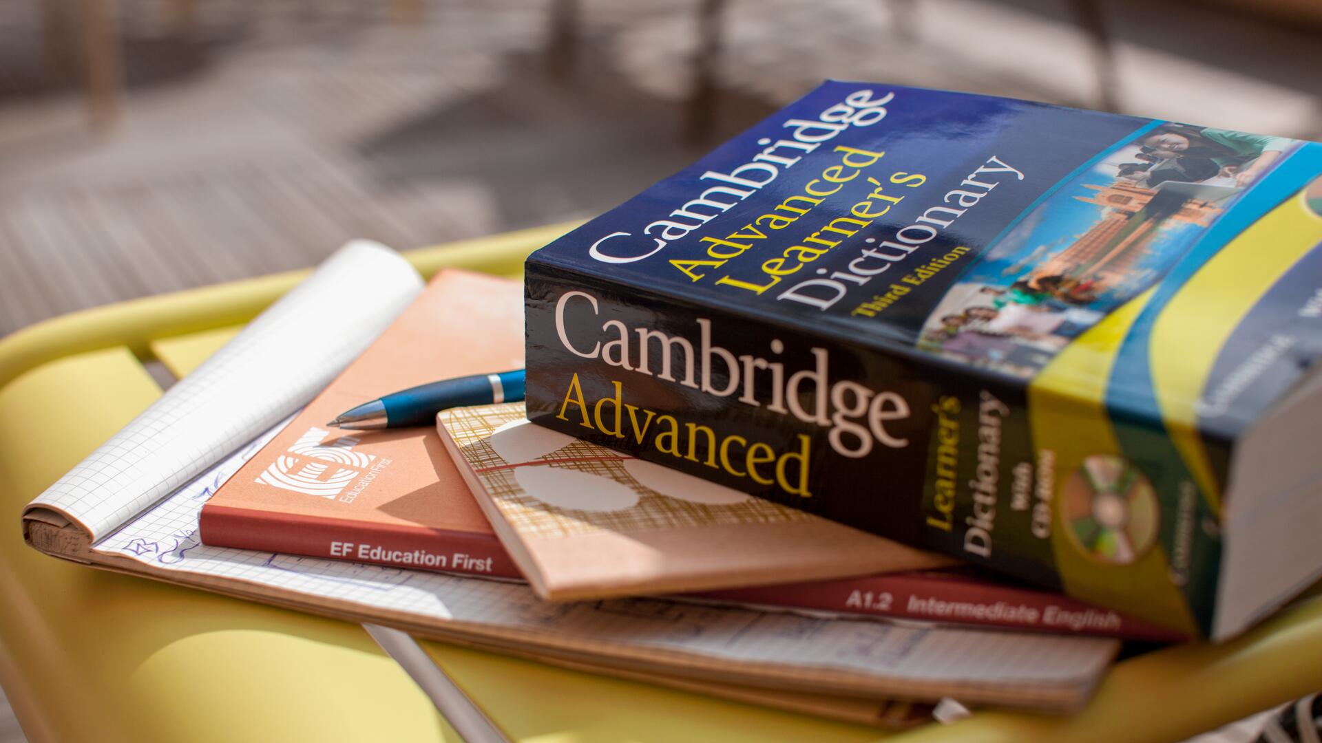 A Cambridge Advanced Learner\'s Dictionary rests on a stack of books and notebooks, with a pen beside it.