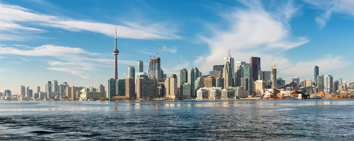 The Toronto skyline, featuring the CN Tower and numerous skyscrapers, is reflected in calm waters under a blue sky with wispy clouds.