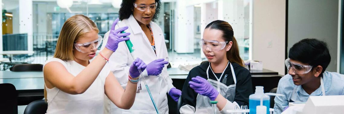 Four students and a teacher are conducting a science experiment in a laboratory, wearing safety goggles and gloves.