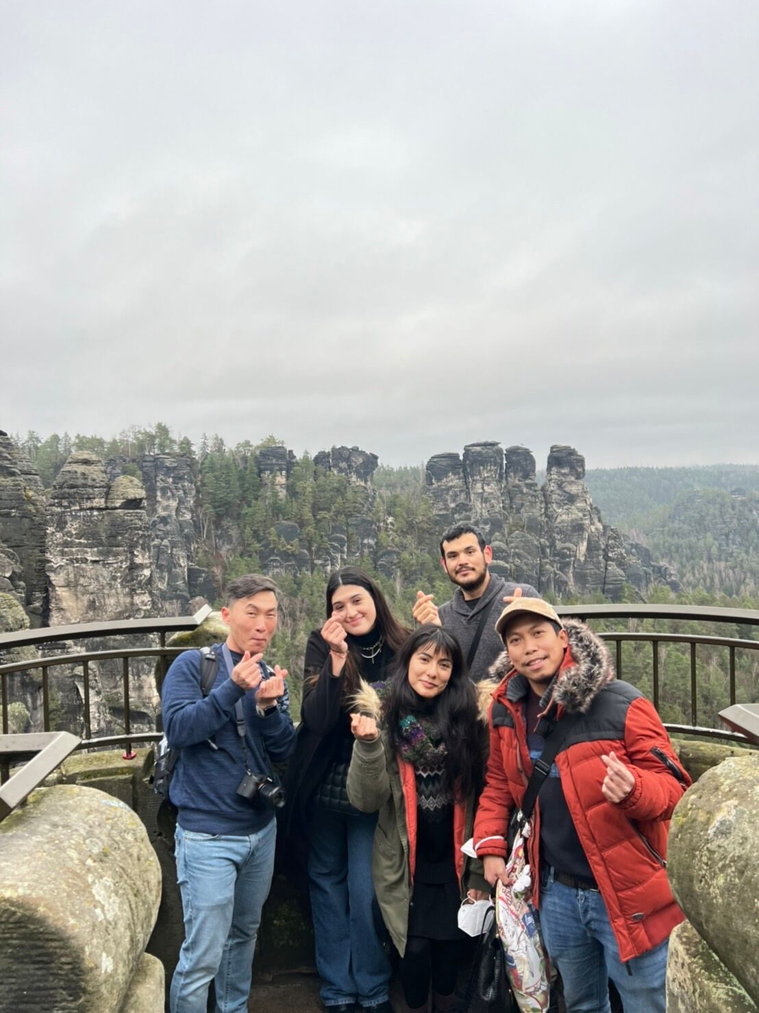A group of five friends pose for a photo on an observation deck, with a stunning rock formation in the background. They are smiling and making heart signs with their hands.