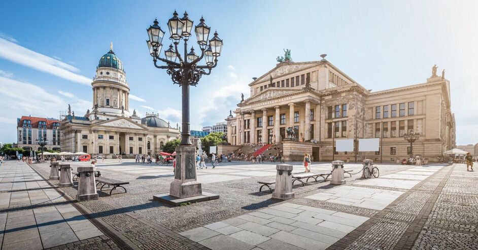 This image shows a wide, sunlit plaza in front of two grand, classical buildings. A prominent street lamp stands in the foreground, with people strolling and benches scattered around the square.