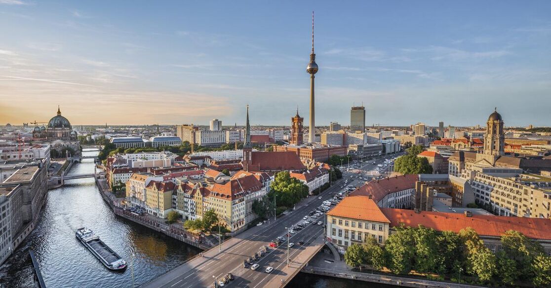 A scenic view of the Berlin skyline featuring the TV Tower, with the Spree River and a barge in the foreground under a clear blue sky.