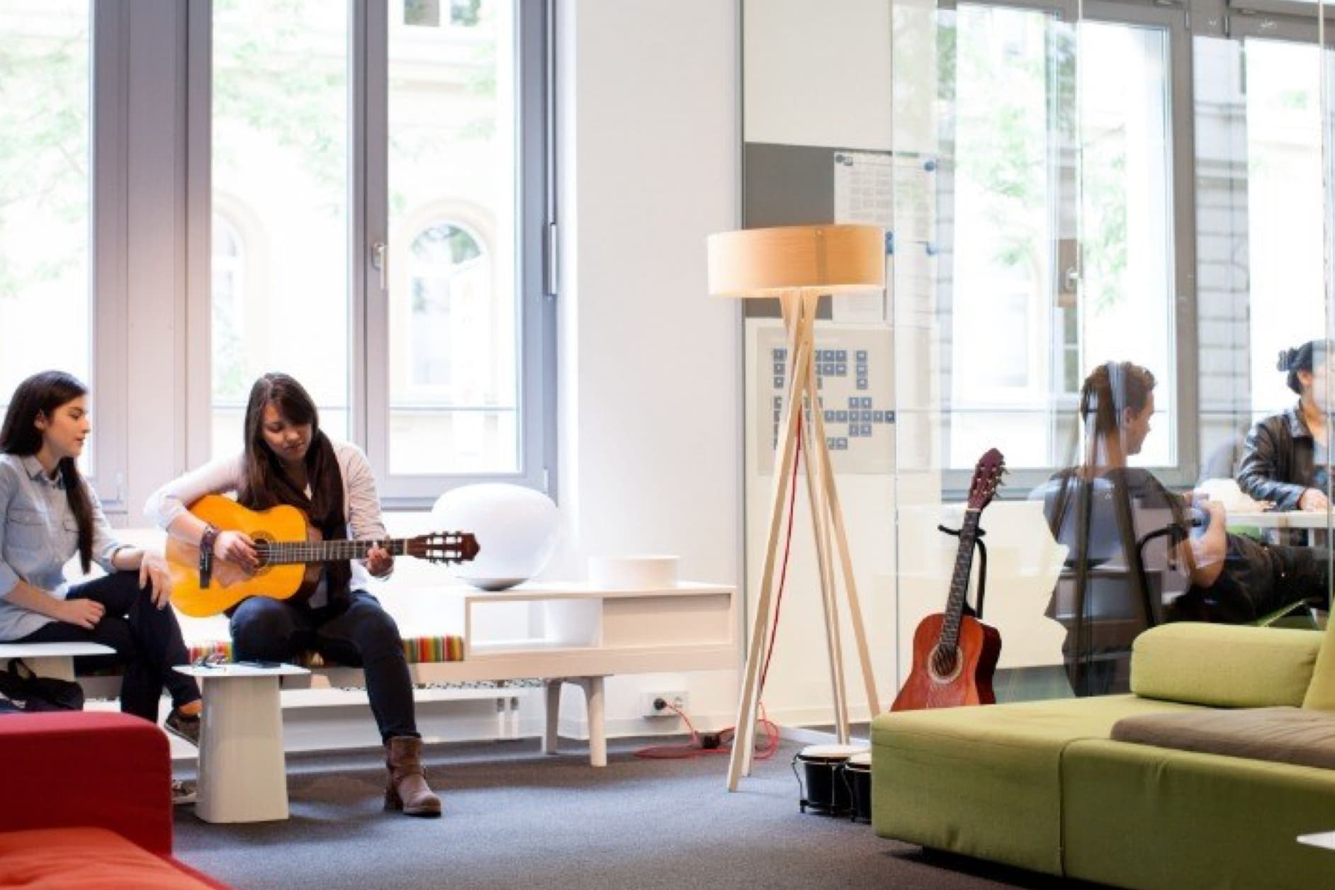 In a bright, modern room, a woman plays an acoustic guitar while two friends listen. Another guitar stands nearby, and a group of people are visible through a glass partition.
