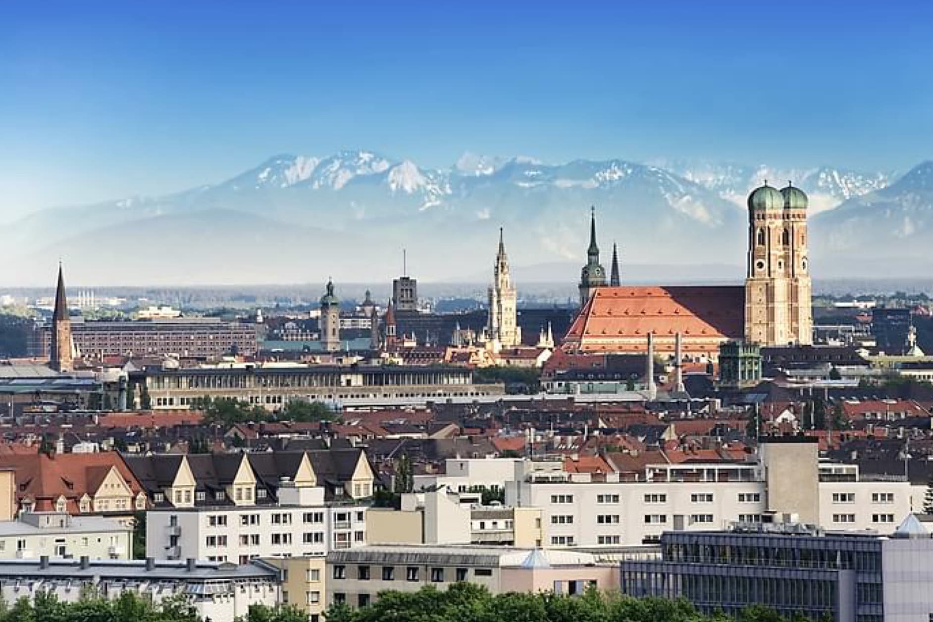 This image shows a panoramic view of the city of Munich, Germany, with its iconic Frauenkirche cathedral and the Alps in the background under a clear blue sky.