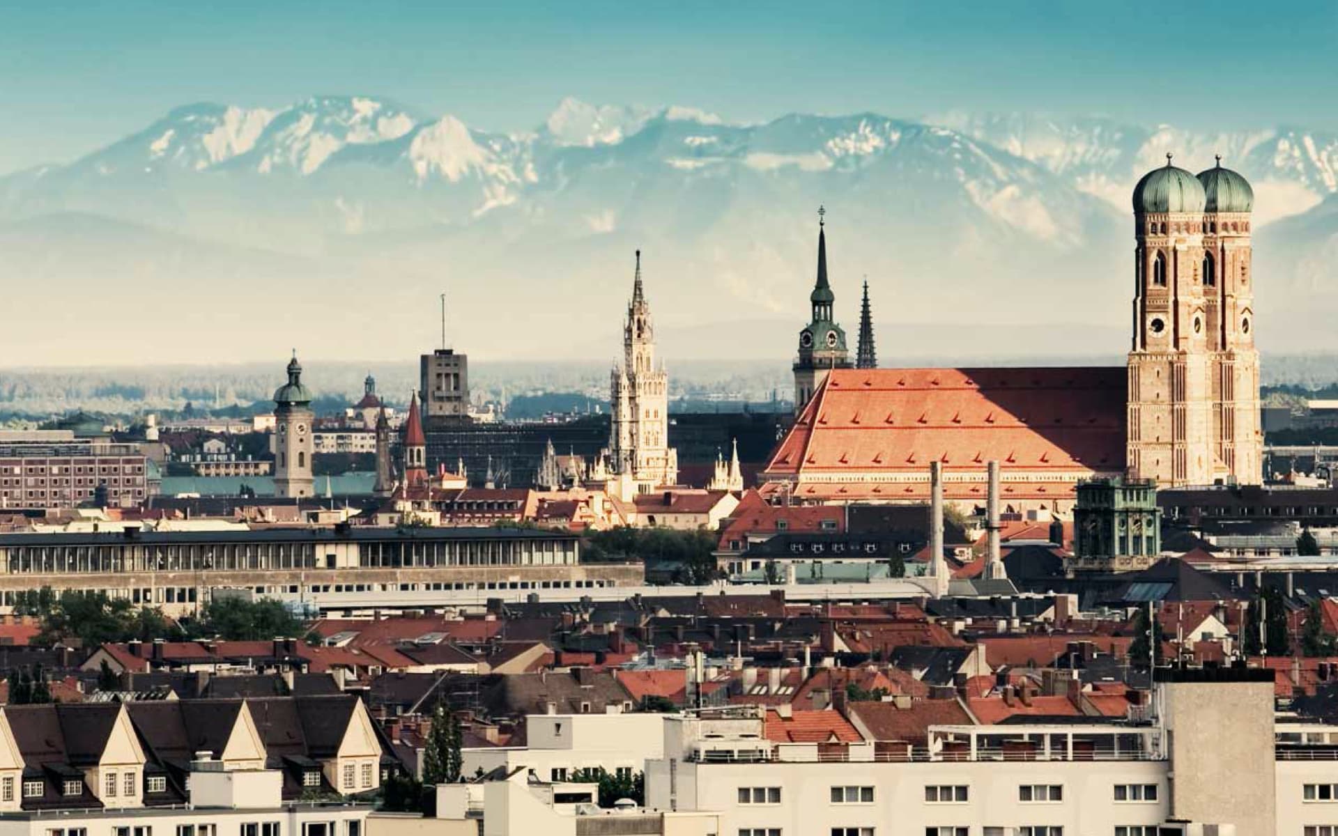 This image captures the cityscape of Munich, with the Alps forming a scenic backdrop. The Frauenkirche, with its distinctive onion domes, is prominently featured.
