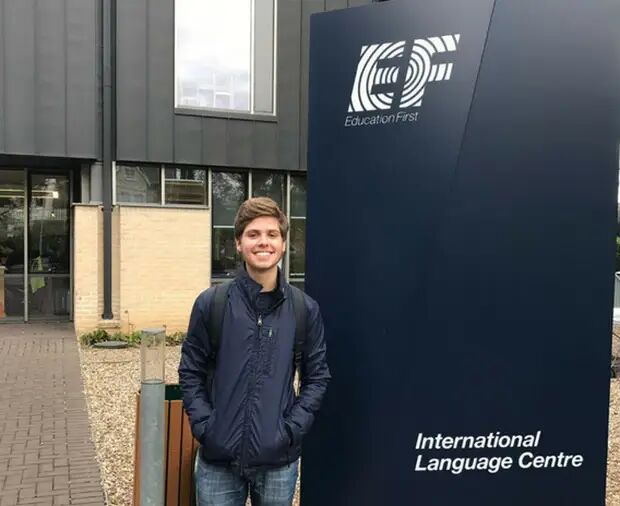 A young man stands in front of the Education First International Language Centre, smiling at the camera. He is wearing a dark jacket and a backpack.