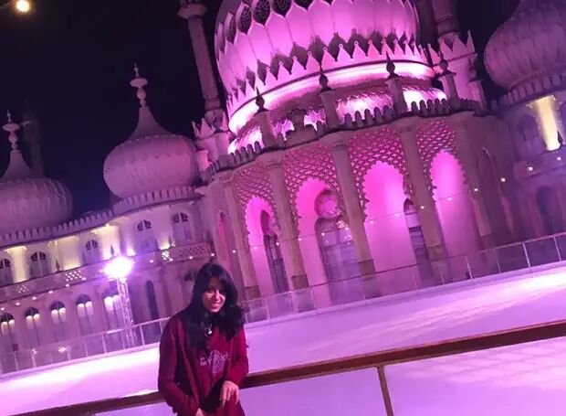 A woman stands on an ice rink in front of the Royal Pavilion in Brighton, illuminated in vibrant pink at night. The architecture is ornate, and the scene feels festive.