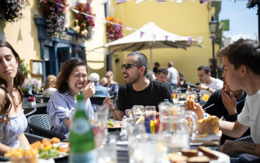 A group of friends are enjoying an outdoor meal on a sunny day, laughing and conversing animatedly.