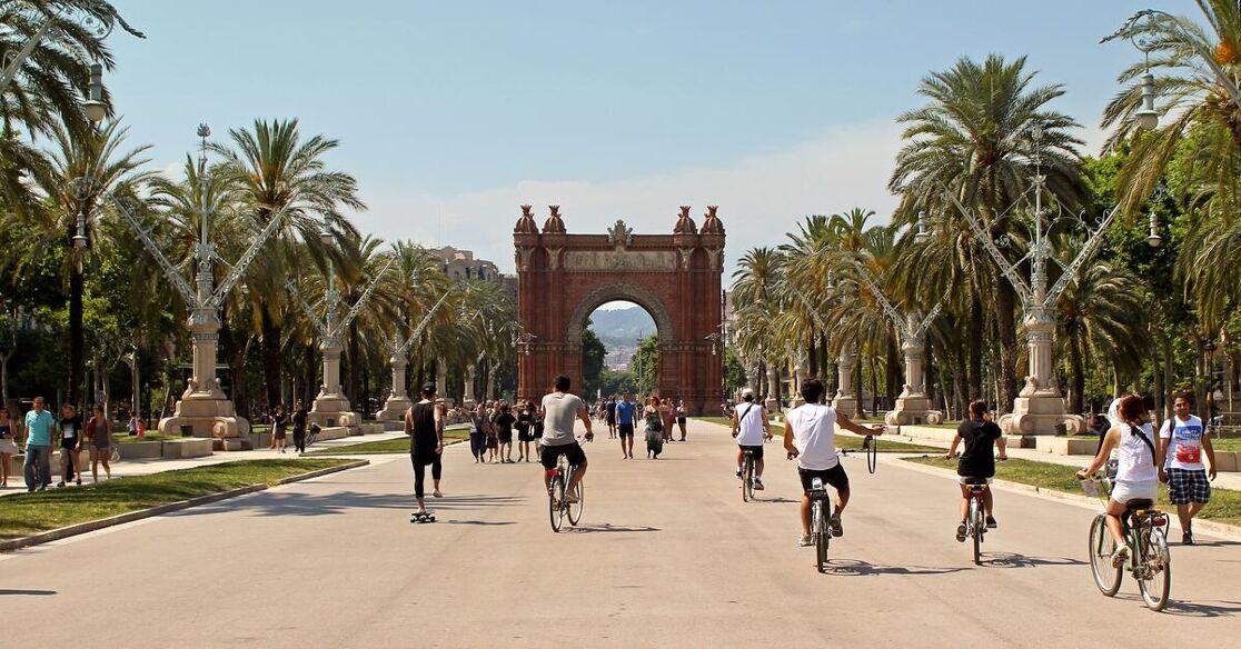 This is a wide, tree-lined avenue leading to the Arc de Triomf in Barcelona, Spain. People are enjoying a sunny day, cycling and walking.