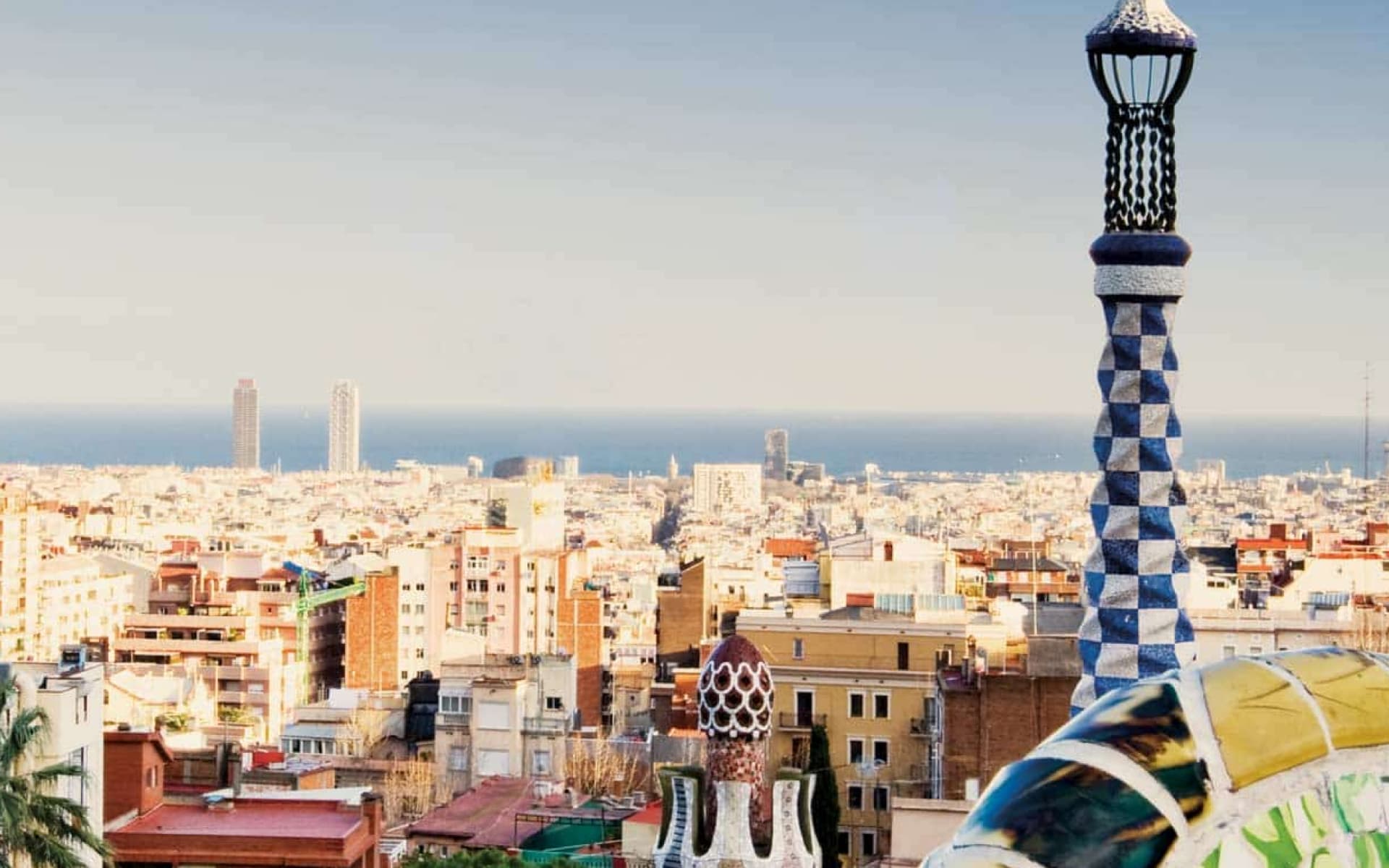A panoramic view of the city of Barcelona, with the sea in the background. Distinctive mosaic structures are visible in the foreground, hinting at Gaudí's architectural style.