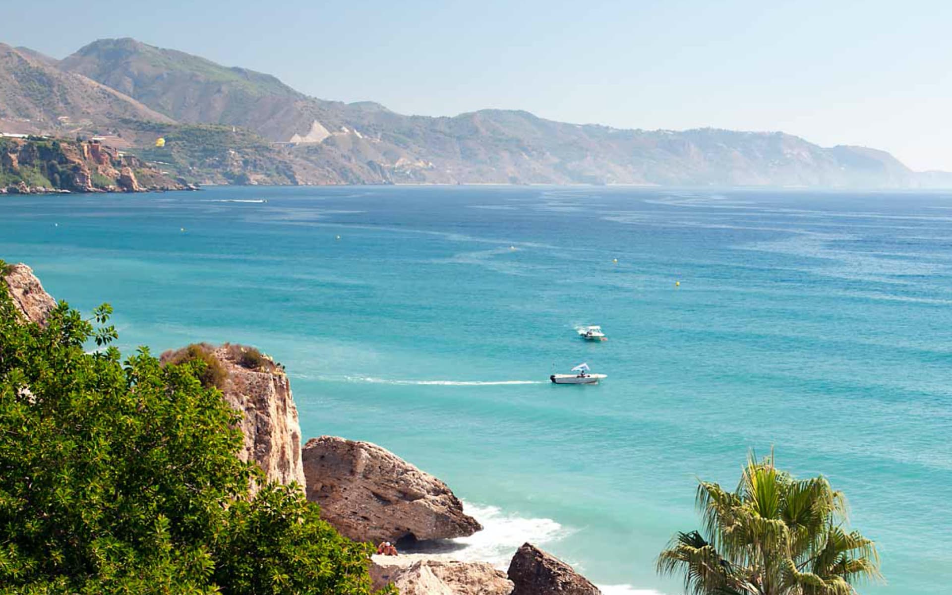 A picturesque view of a coastal landscape featuring turquoise waters, rocky cliffs, and mountains in the background. Two boats glide across the calm sea, offering a serene scene.