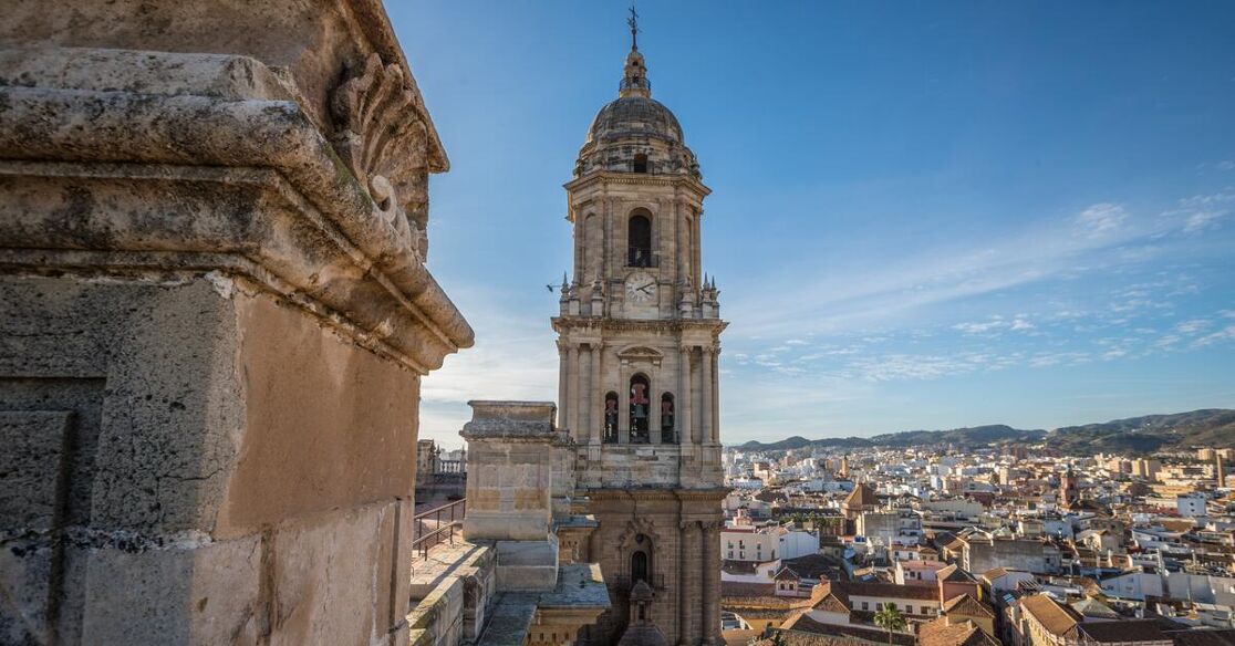 A tall bell tower dominates the skyline of a city, with mountains visible in the distance.