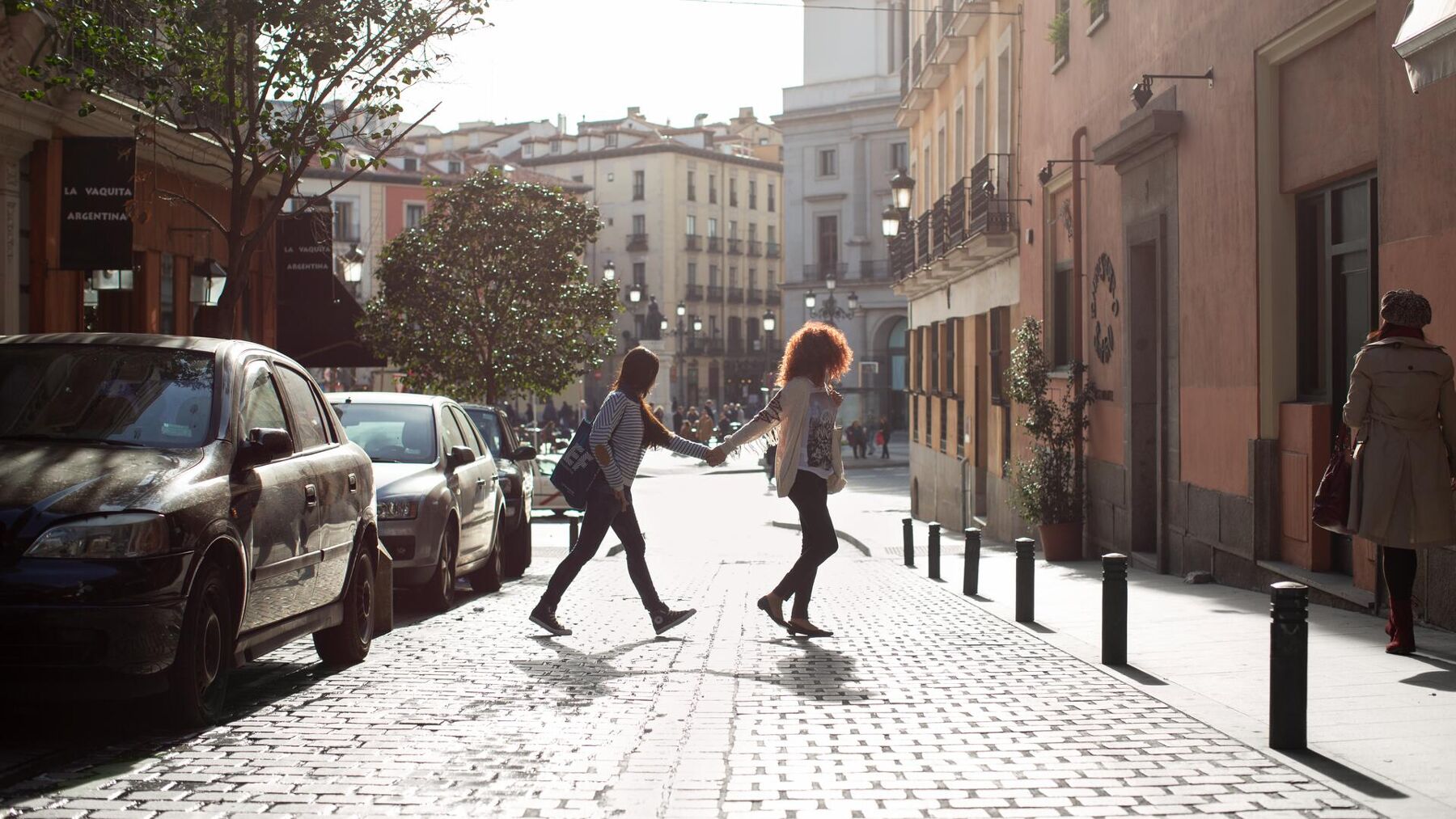 Two people are crossing a cobblestone street in a European city, holding hands as they walk across a crosswalk. Cars are parked along the side of the street, and buildings line the opposite side.