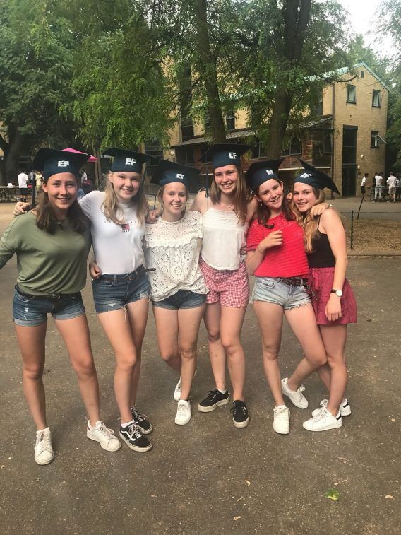 Six young women wearing graduation caps pose for a photo outdoors. They are dressed casually in shorts and t-shirts, smiling and looking at the camera.