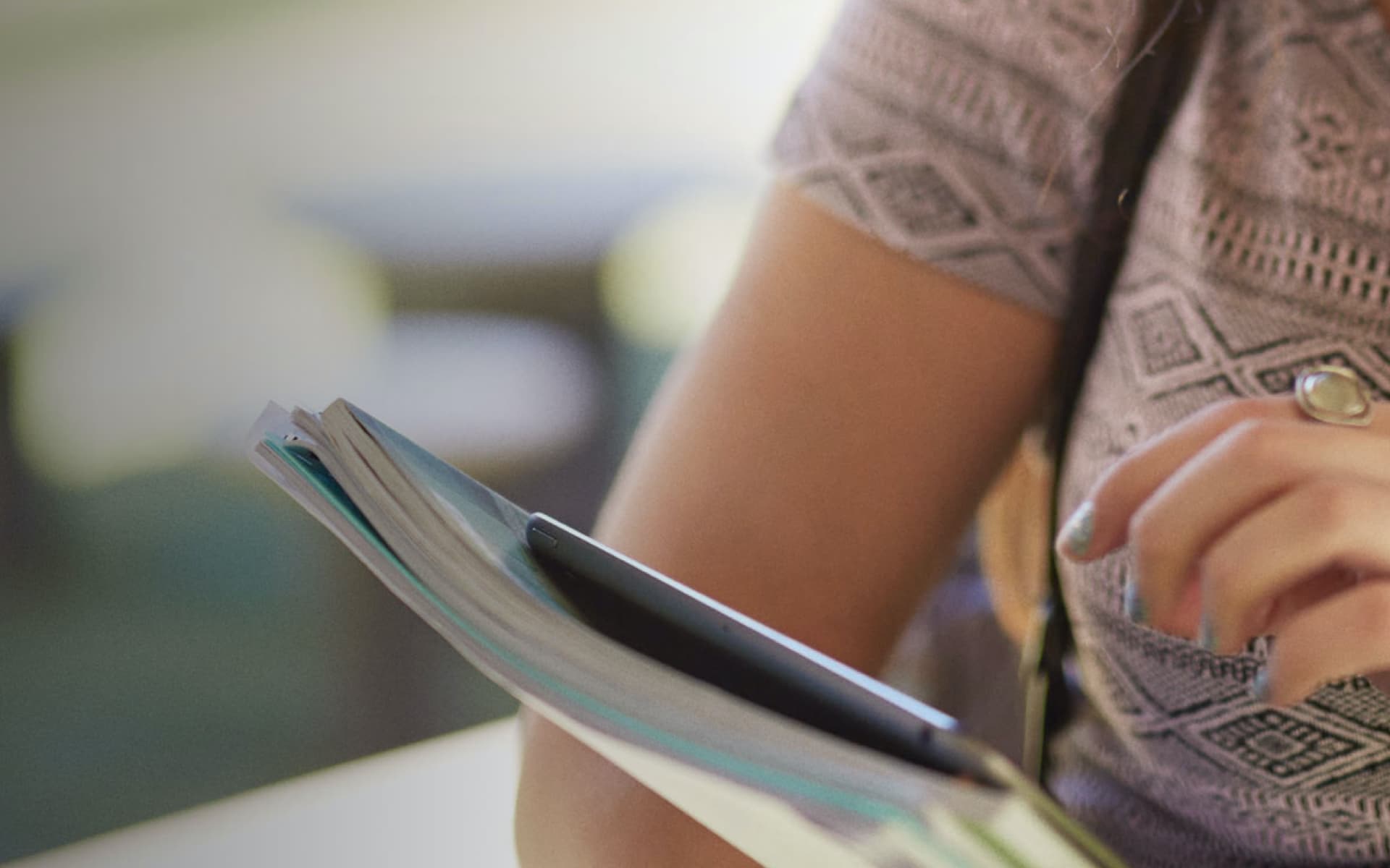 A person is reading a booklet or book, which is open with a pen tucked inside. The focus is on the hand and the booklet.