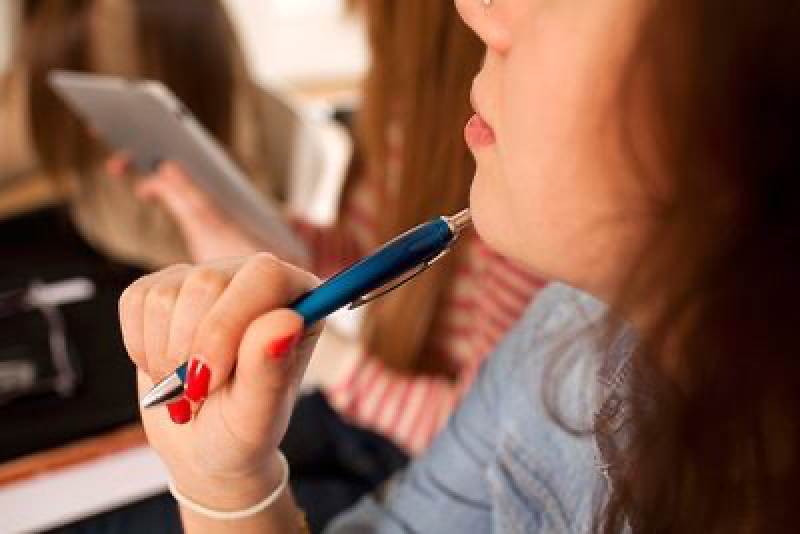 A young woman thoughtfully chews on a pen while sitting near a tablet, appearing to be studying.