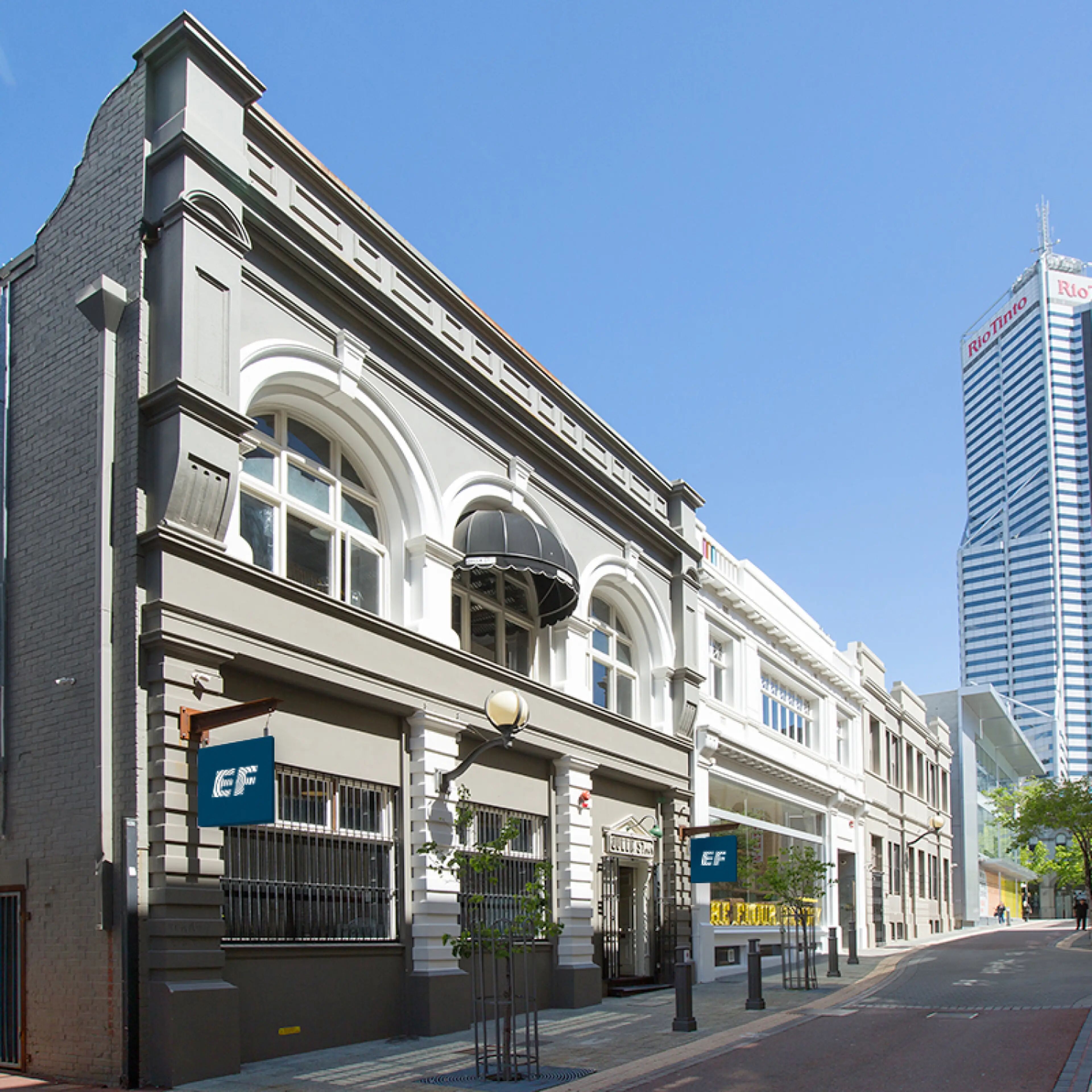 The image shows a row of historic buildings with ornate architecture, one of which features an "EF" logo, suggesting it might be an educational institution. A modern skyscraper looms in the background.