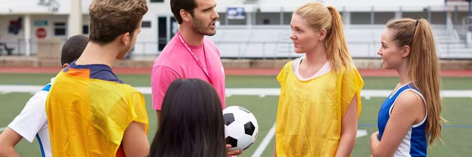 A coach is talking to a soccer team on the field. They are wearing training bibs and appear to be listening to instructions.
