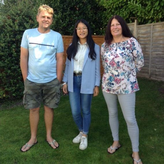 Three people are standing on a lawn in front of a wooden fence and bushes. A man is on the left, a young woman in the middle, and a woman on the right.