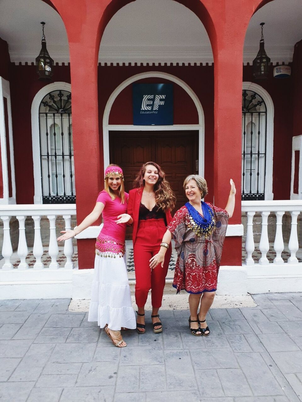 Three women are posing in front of a red building with an "EF" logo. They are dressed in colorful outfits and appear to be enjoying themselves.