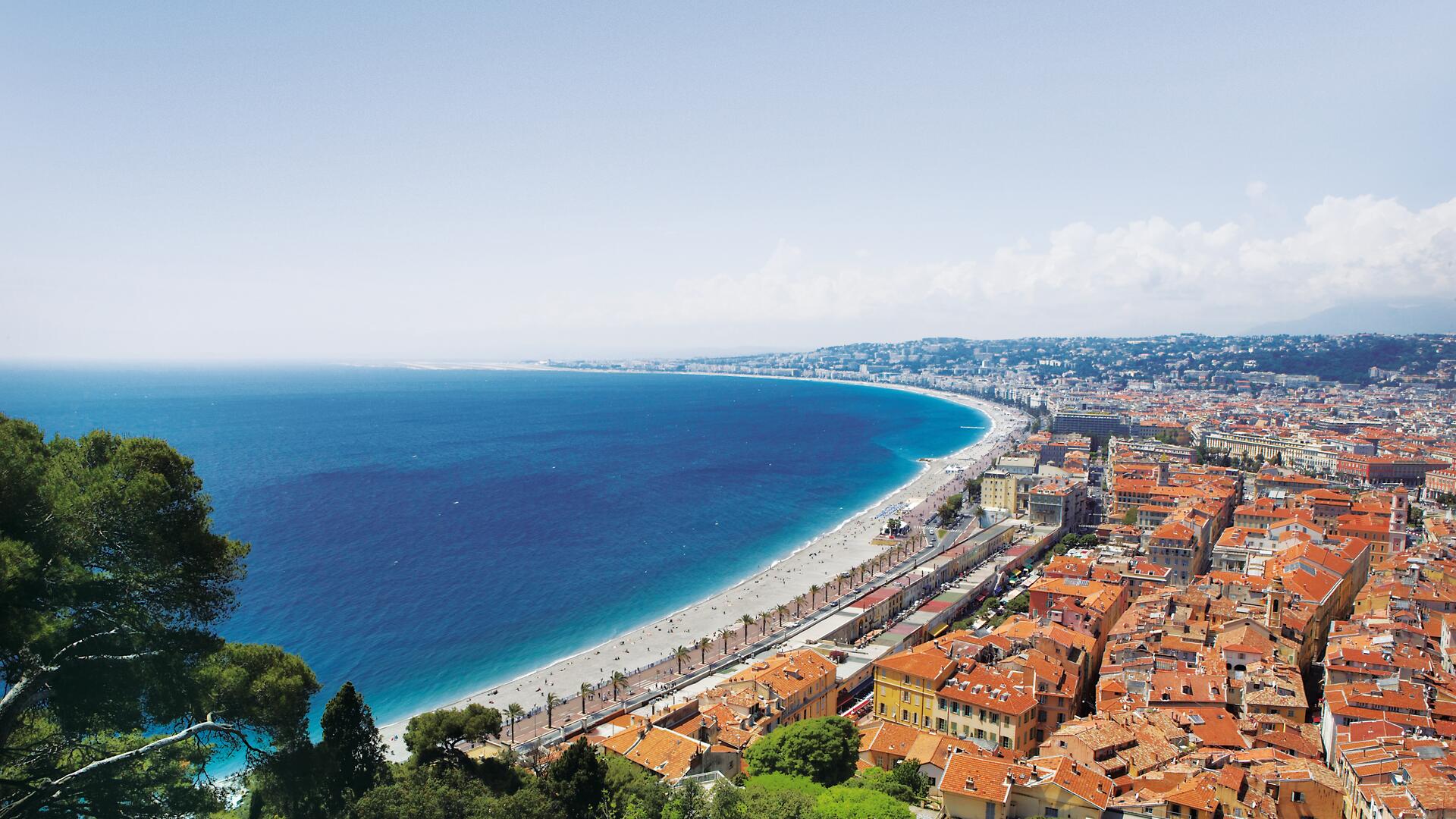 This is a scenic view of the Nice coastline in France, featuring a long pebble beach, the blue Mediterranean Sea, and the cityscape with its characteristic red-tiled roofs.