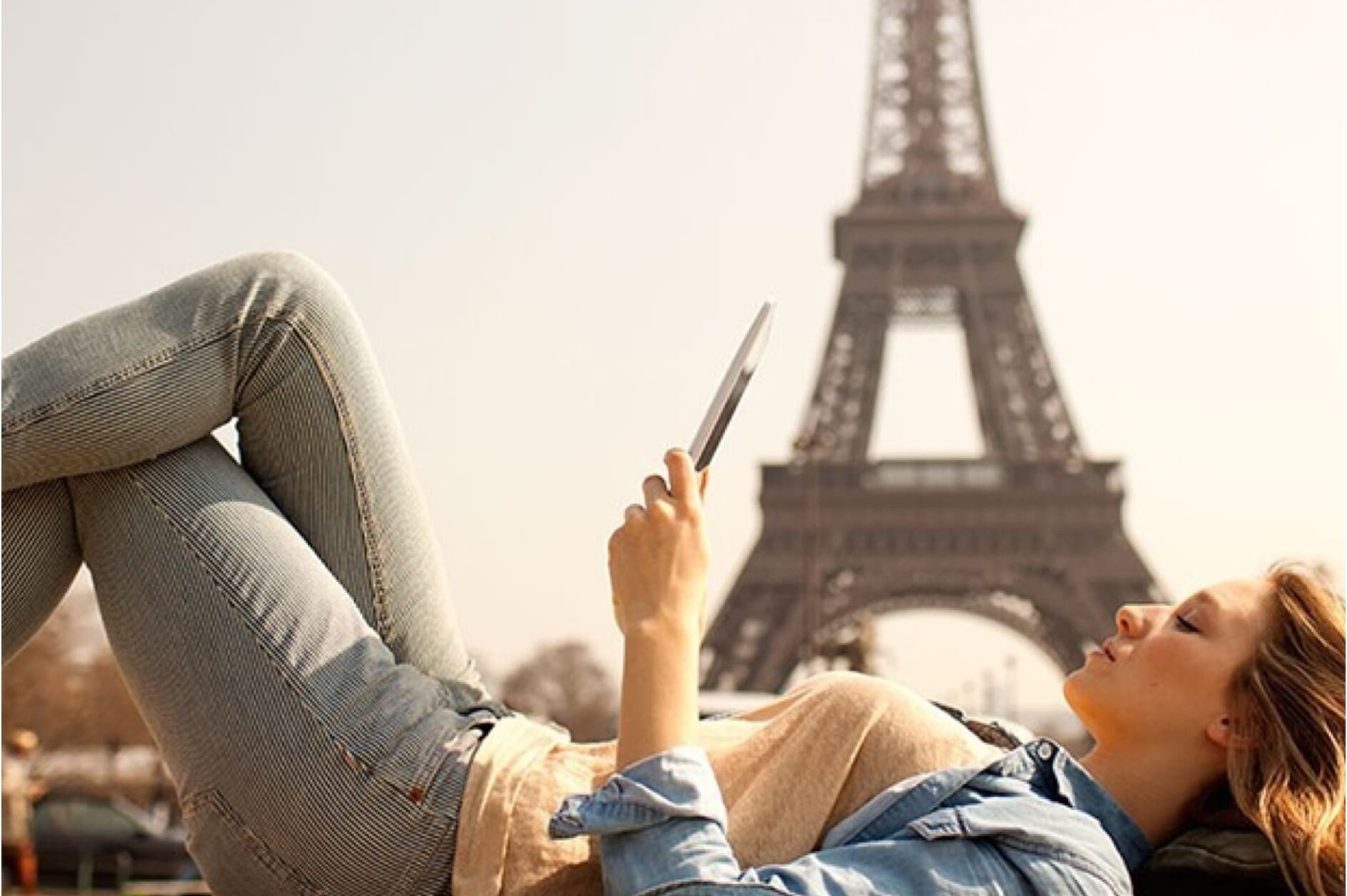 A young woman is relaxing outdoors, lying on her back and looking at her phone, with the Eiffel Tower visible in the background.