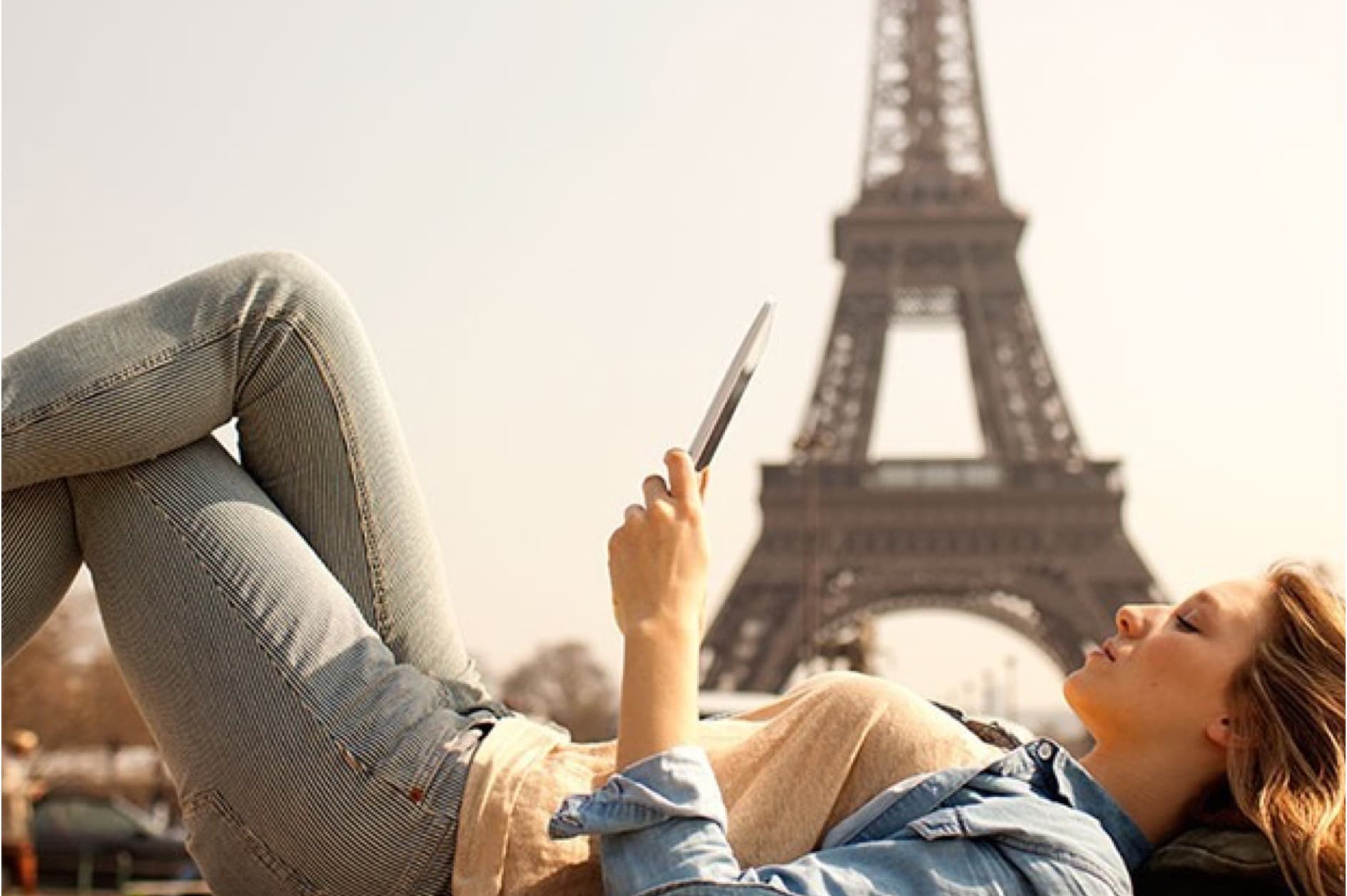 A young woman is relaxing outdoors, lying on her back and looking at her phone, with the Eiffel Tower visible in the background.