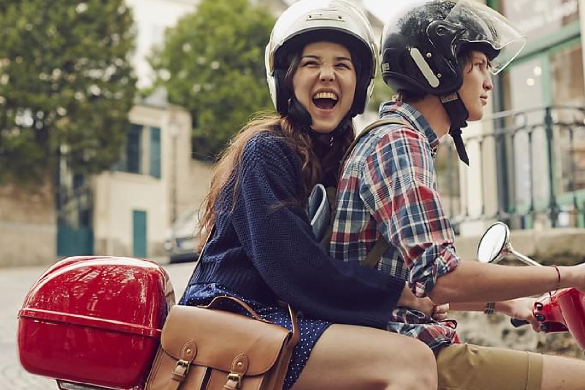 A young couple rides on a scooter through a city, both wearing helmets and appearing to have a great time.