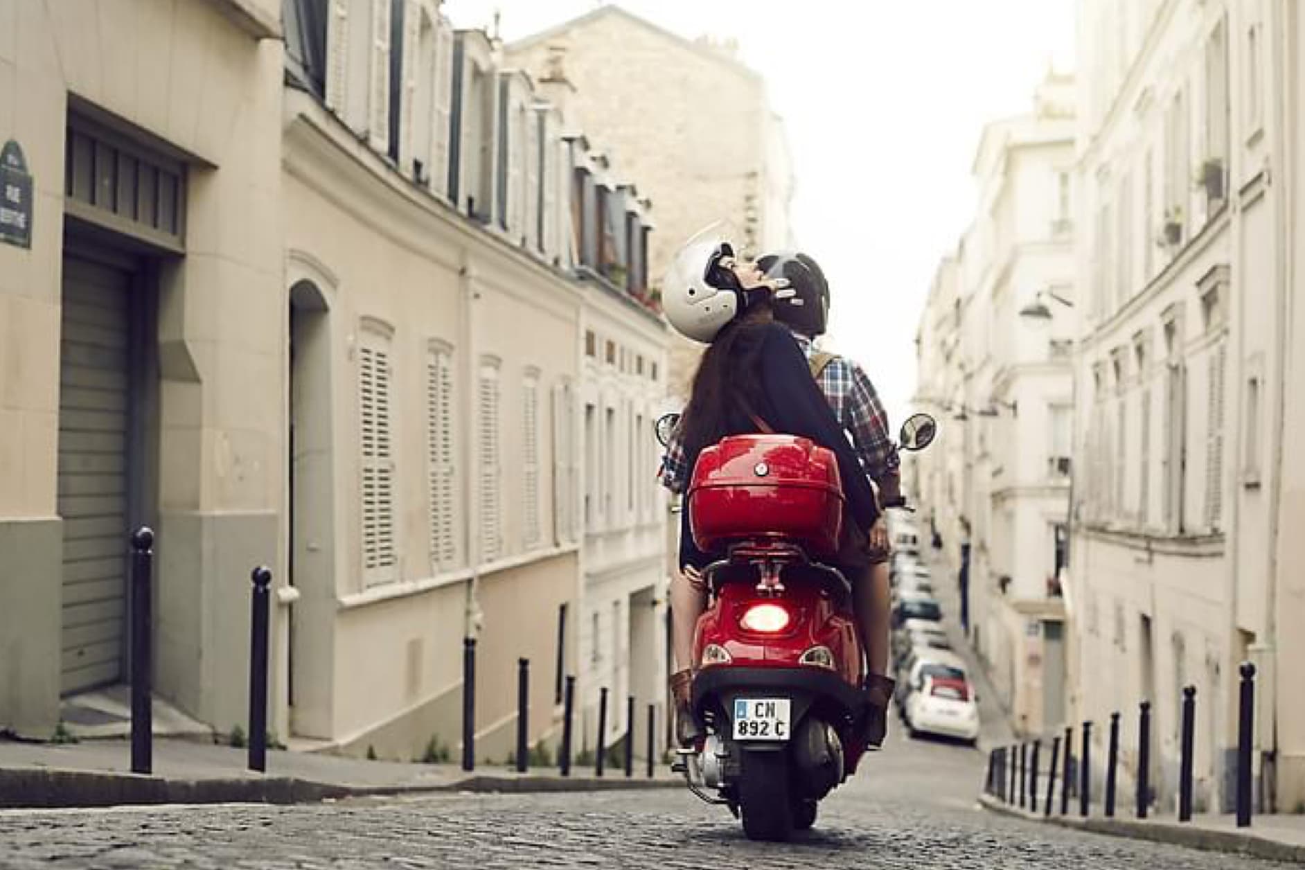 A couple rides a red scooter up a cobblestone street in a European city, with charming buildings lining both sides.