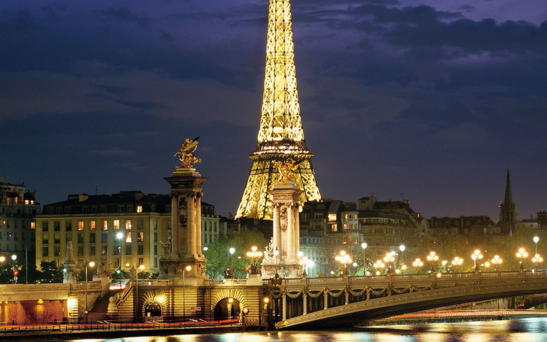The image shows the Eiffel Tower in Paris at night, illuminated and majestic, with the Seine River and a bridge in the foreground, creating a romantic and iconic cityscape.