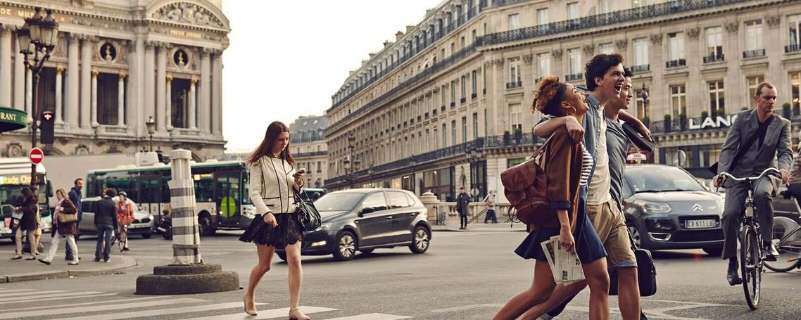 A group of friends crosses a busy Parisian street, with the magnificent Opera House in the background and cars passing by.