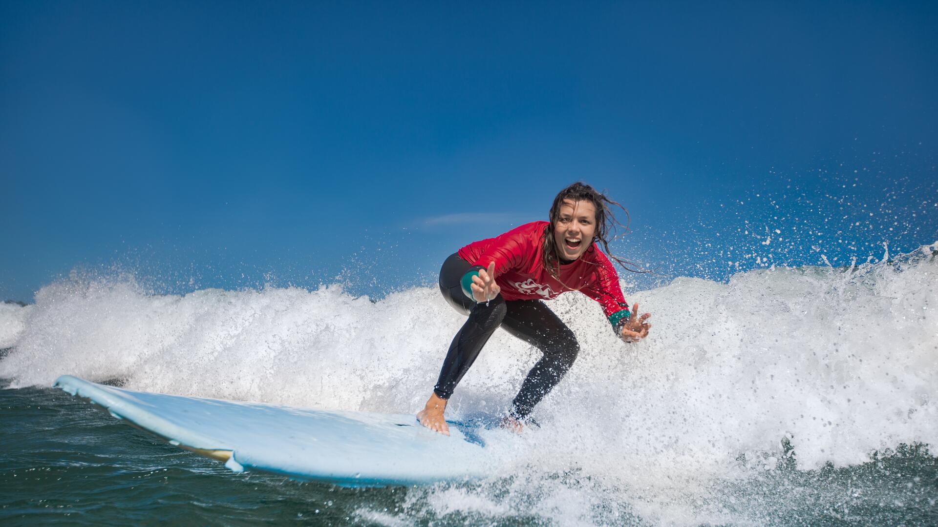 A woman in a red rash guard and black wetsuit surfs a wave on a blue surfboard, with a big smile and arms outstretched, enjoying the ride.