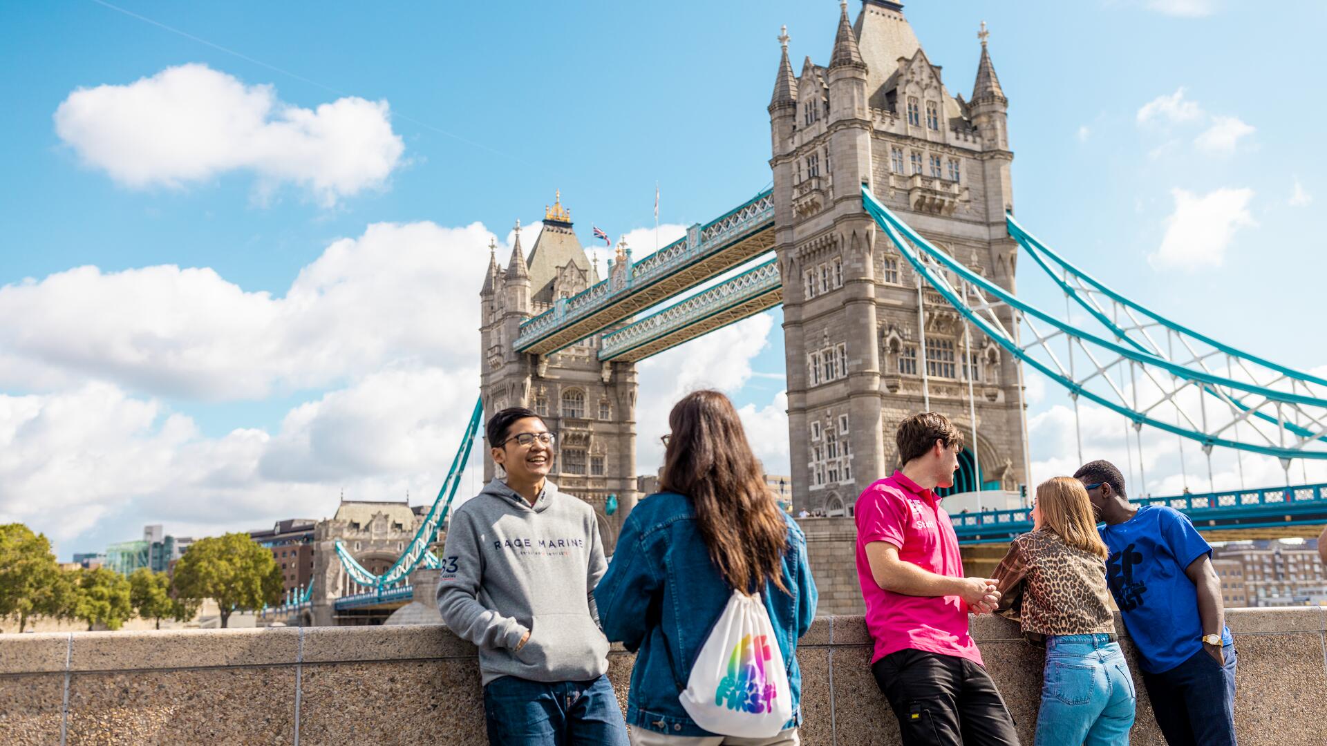 Four young people are standing in front of Tower Bridge in London, enjoying a sunny day. The famous landmark bridge is clearly visible in the background.