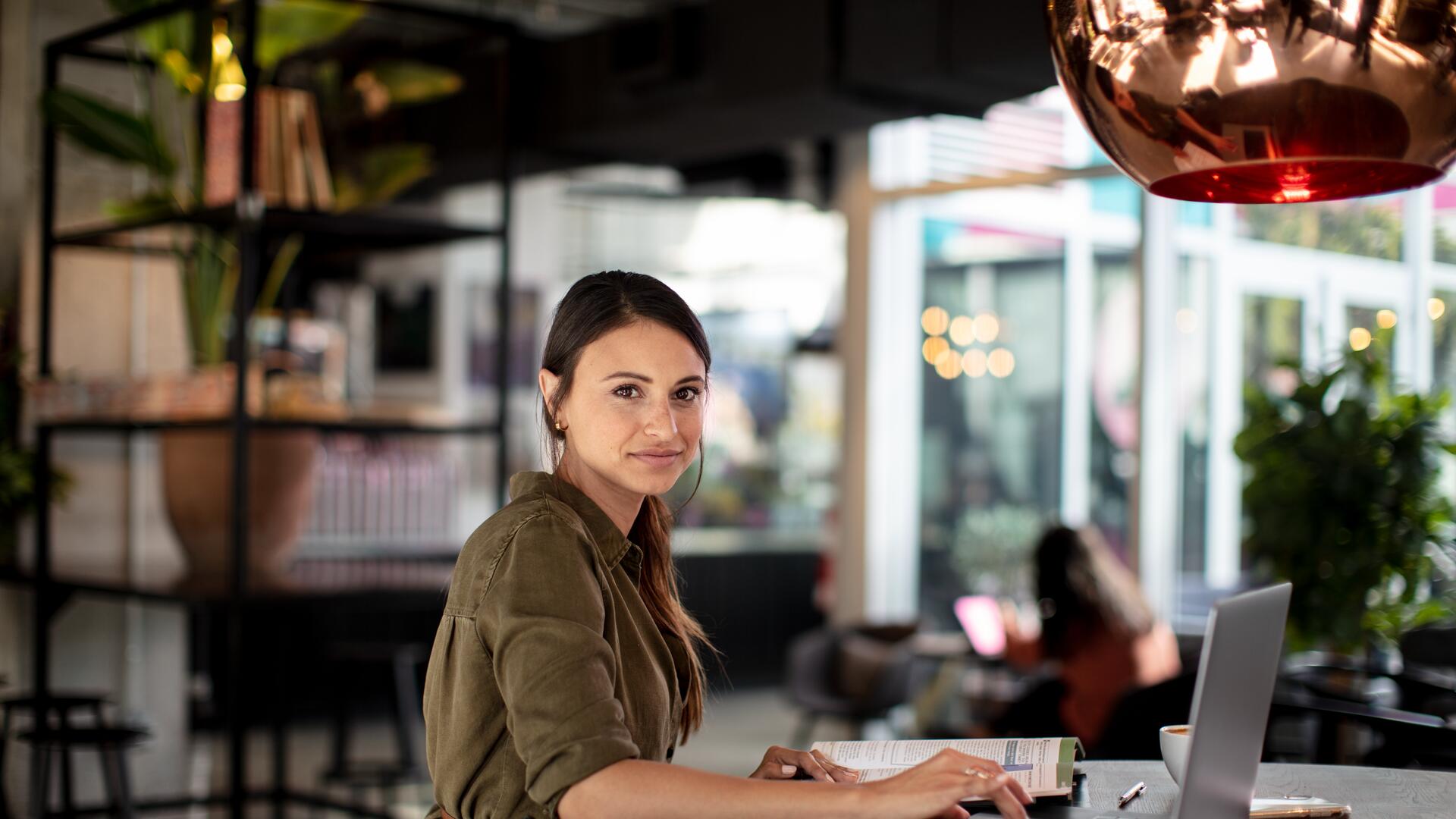 A woman sits at a table with a laptop, looking towards the camera. She is in a bright, modern cafe or co-working space.