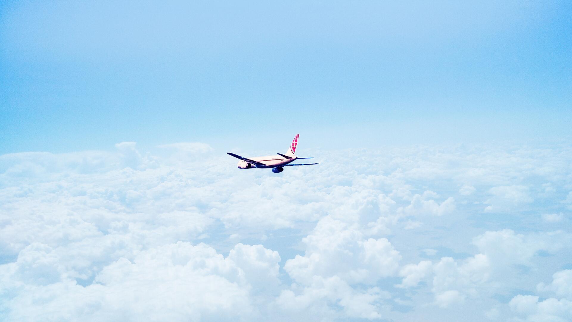 An airplane is seen soaring high above a blanket of clouds, with a blue sky overhead. The plane's wings are extended as it glides through the air.
