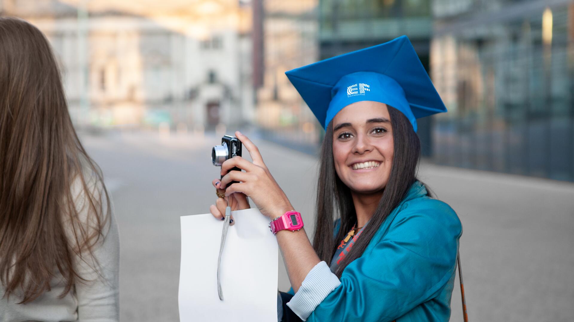 A young woman in a blue graduation outfit and cap holds a camera and a white bag, smiling. She appears to be capturing her special day.