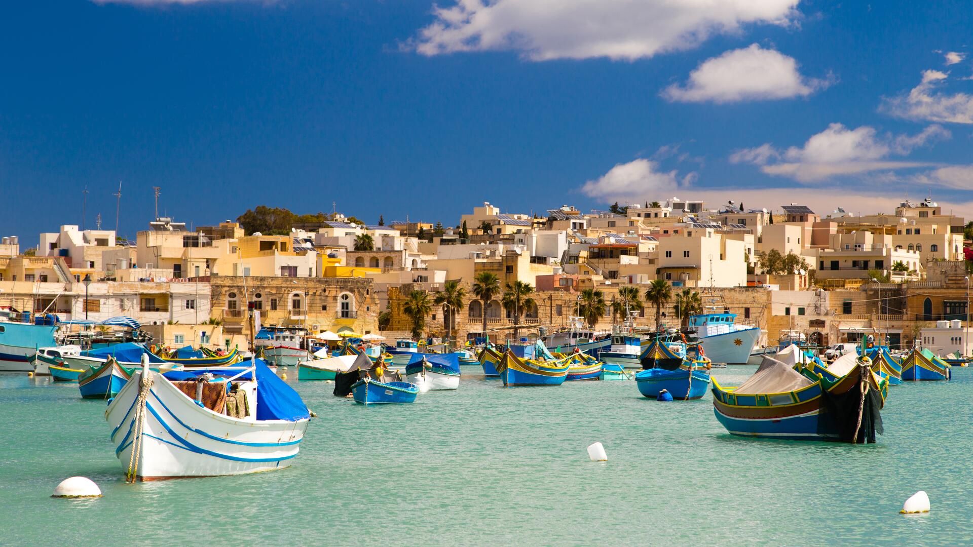 This image depicts a picturesque harbor filled with traditional colorful fishing boats, likely in a Mediterranean coastal town. The buildings in the background are clustered together, creating a charming waterfront scene under a clear blue sky.