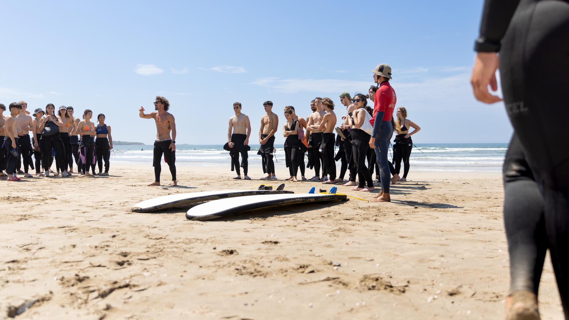 A group of people in wetsuits are gathered on a sandy beach, listening to an instructor. Two surfboards lie on the sand in the foreground, suggesting a surfing lesson or activity.