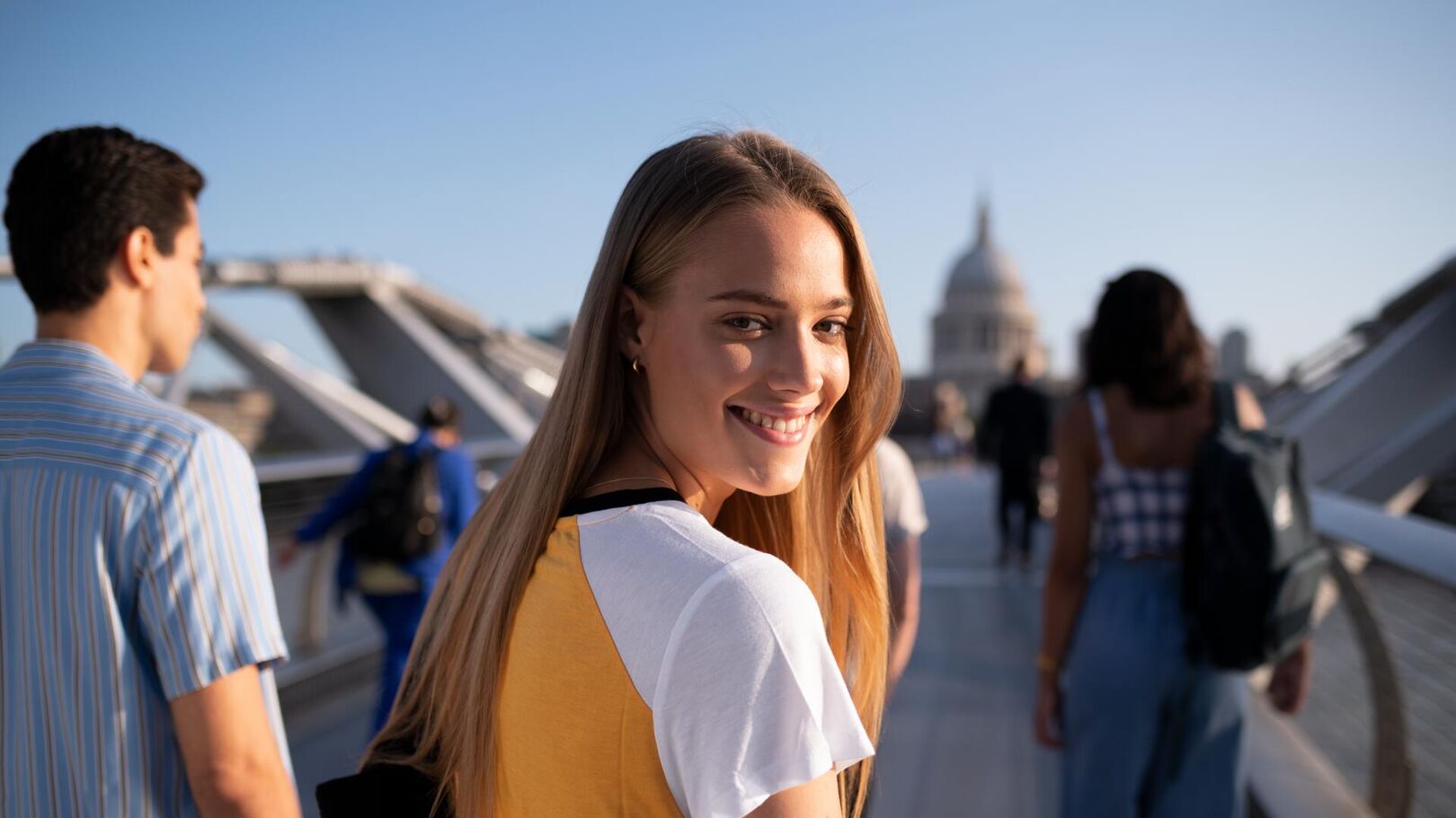 A young woman with long blonde hair smiles over her shoulder as she walks across a bridge. Other people and a domed church are visible in the background.