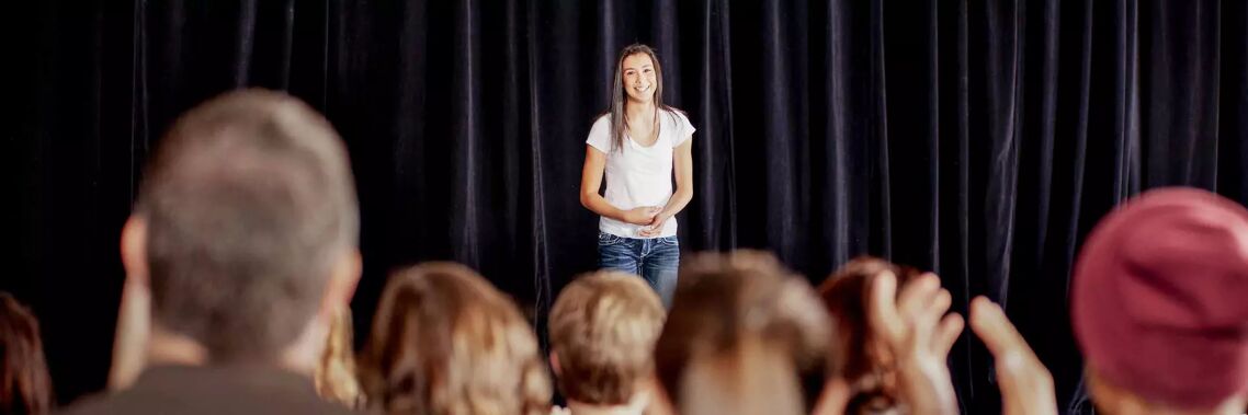 A young woman stands on a stage in front of a black curtain, speaking to an audience. She is wearing a white t-shirt and jeans.