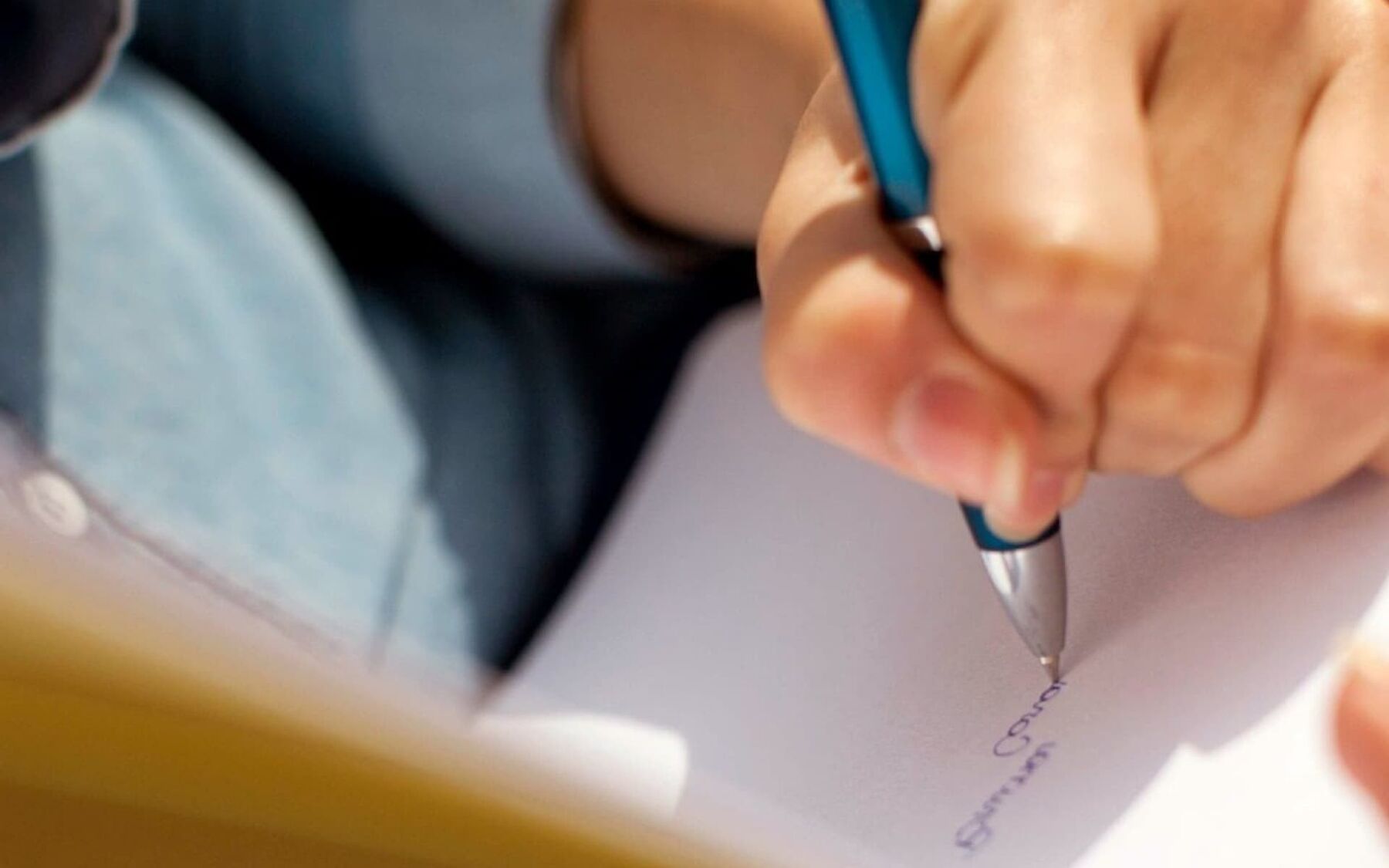 A close-up shot focuses on a hand holding a blue pen and writing on a piece of paper. The image captures the act of jotting down a note or signing a document.