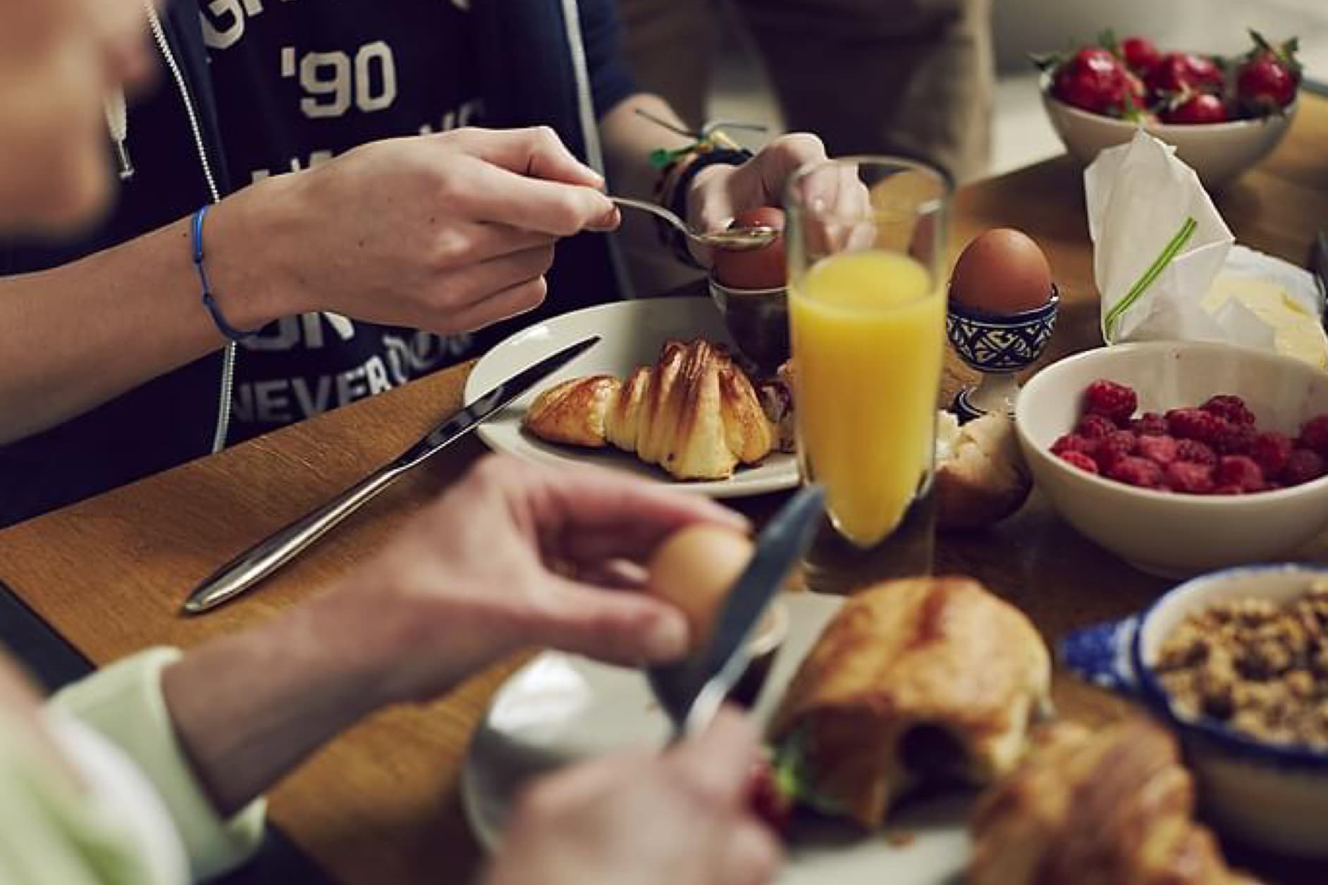 This image shows a group of people enjoying a delicious breakfast, complete with croissants, boiled eggs, fresh berries, and orange juice, set on a wooden table.