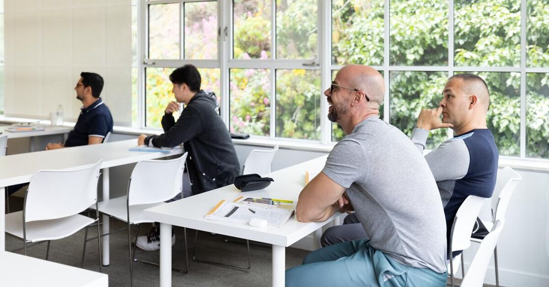 Four men are seated at tables in a bright classroom with large windows overlooking trees. They appear to be listening to a presentation.