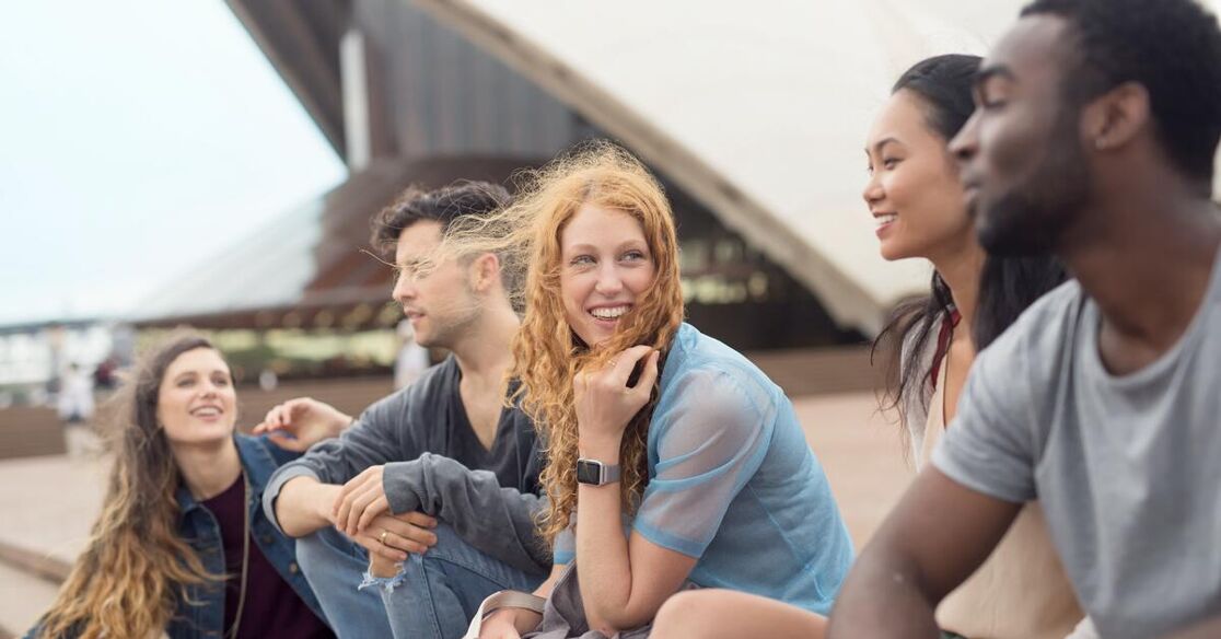A diverse group of young adults are sitting together outdoors, appearing to be in good spirits and enjoying each other's company. The background features a modern architectural structure.