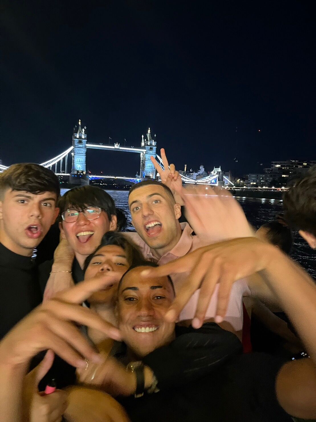 A group of five young men are taking a selfie at night, with the illuminated Tower Bridge in the background. They are smiling and posing for the camera.