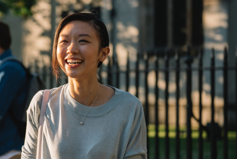 A young Asian woman is smiling brightly as she walks outside on a sunny day. She is wearing a gray sweatshirt and a necklace with a pearl.