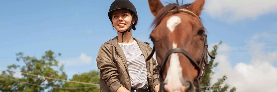 A young woman wearing a helmet and a jacket is riding a brown horse. She is smiling at the camera, and the sky is blue with some clouds.
